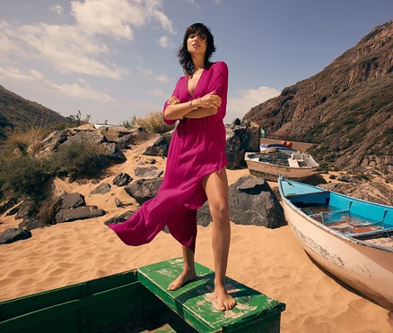 Eine Frau mit dunklen Haaren steht barfuß in einem pinken Kleid auf einem grünen Boot am Strand.