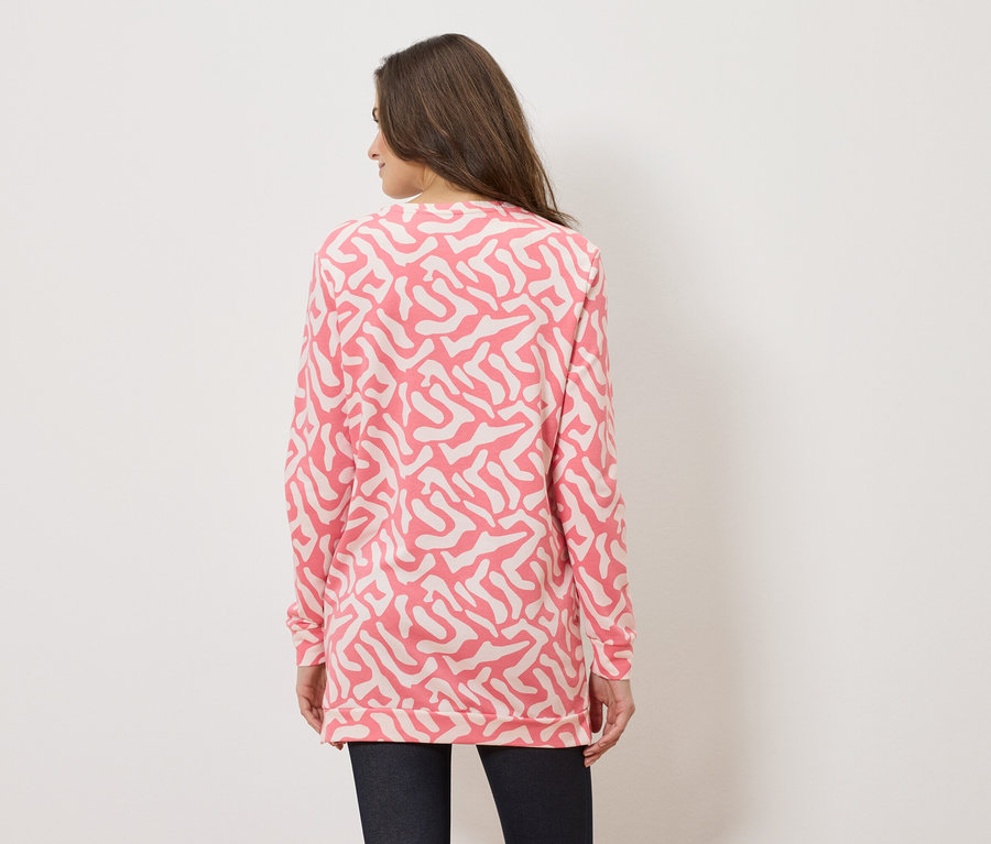 Rückansicht einer Frau mit langen braunen Haaren, die einen rosa Pullover mit abstraktem weißem Muster und dunkle Hosen trägt.