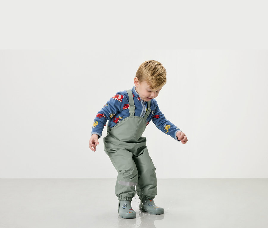 Ein kleiner Junge spielt in grüner Regenhose und Gummistiefeln mit Wasser.