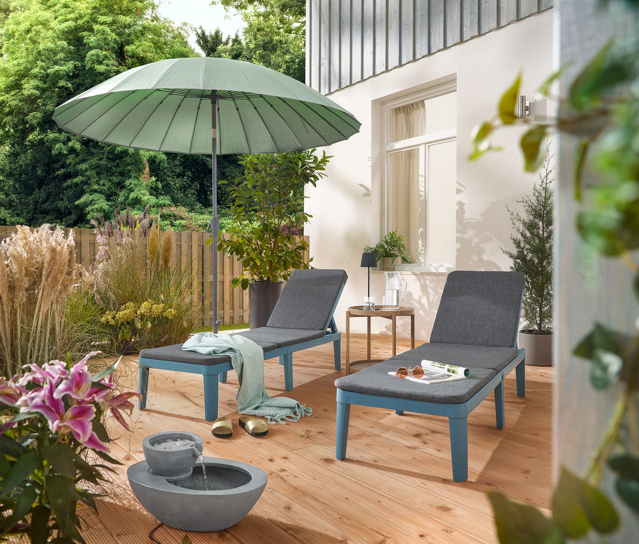 Auf einer Terrasse stehen zwei Liegen unter einem hellgrauen Leco Shanghai Sonnenschirm und neben einem Solar-Gartenbrunnen.