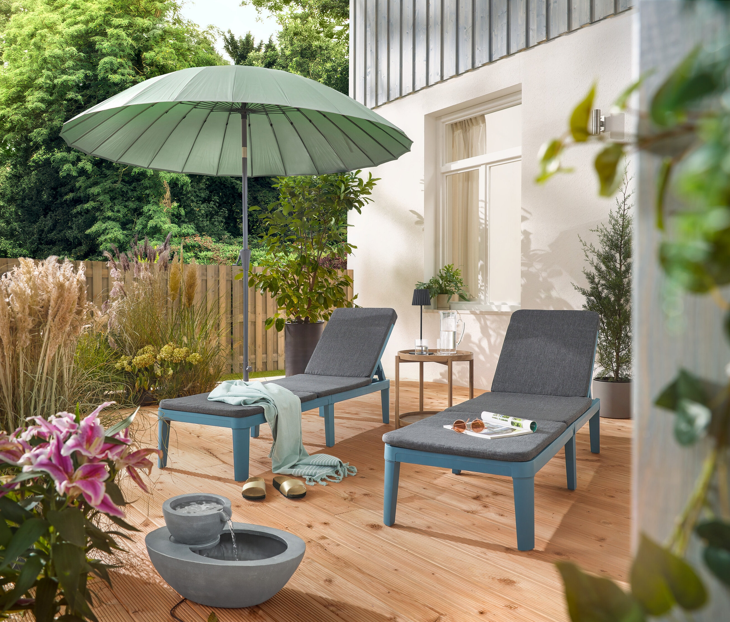 Auf einer Terrasse stehen zwei Liegen unter einem hellgrauen Leco Shanghai Sonnenschirm und neben einem Solar-Gartenbrunnen.