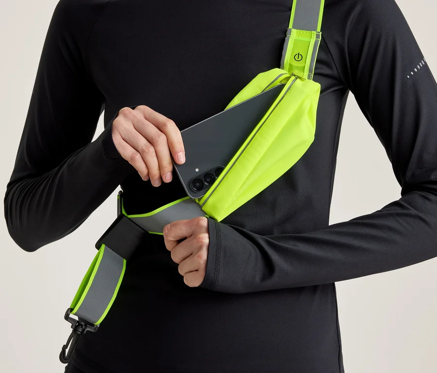 Person in schwarzem Langarmshirt steckt ein schwarzes Smartphone in eine neongelbe Umhängetasche mit reflektierendem Detail.
