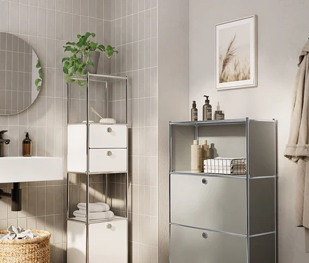 Zwei Metall-Möbelstücke in einem modernen Badezimmer. Links steht ein weißes Metall-Regal »CN3 Slim Line« mit 2 Schubladen und 1 Klappe, darauf eine Pflanze und Handtücher. Rechts eine hellgraue Metall-Kommode »CN3 Slim Line« mit 2 Klappenfächern und einer offenen Ablage mit Flaschen.