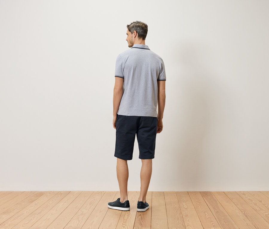 Mann von hinten, trägt ein graues Polohemd, kurze blaue Hosen und schwarze Schuhe.