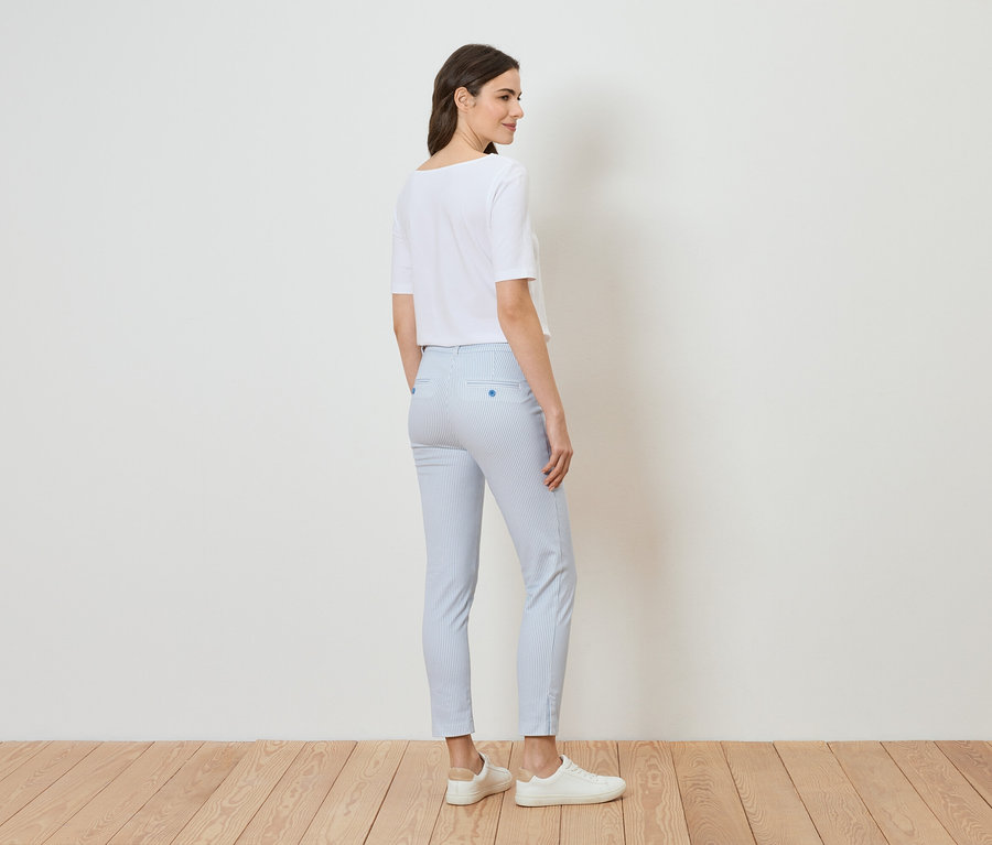 Eine Frau steht in einem hellen Raum, trägt ein weißes T-Shirt, hellblau gestreifte Hosen und weiße Schuhe.