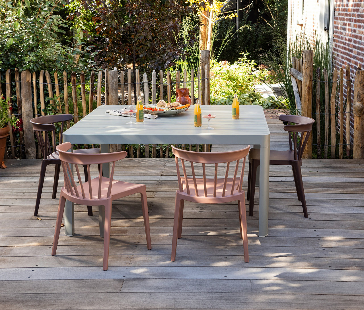 Auf einer Terrasse stehen ein Tisch mit Speisen und Getränken und vier Stühle, darunter zwei rosafarbene WOOOD Gartenstühle.