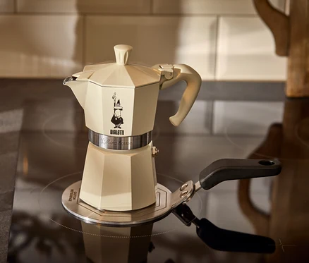 Ein cremefarbener Espressokocher Bialetti Moka Express 190 ml steht auf einer Bialetti Adapterplatte Induktion 13 cm neben einem Glas Espresso.