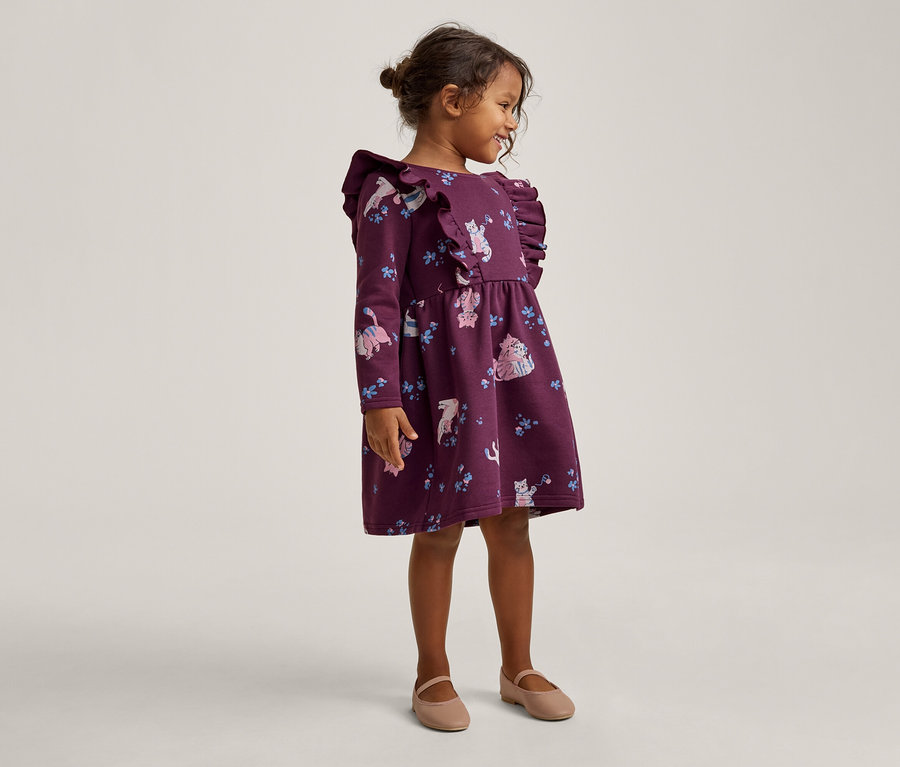 Ein junges Mädchen steht lächelnd und blickt nach rechts, trägt ein dunkelviolettes Kleid mit langen Ärmeln, Rüschen und einem Muster aus Katzen und Blumen, dazu beigefarbene Ballerinas.