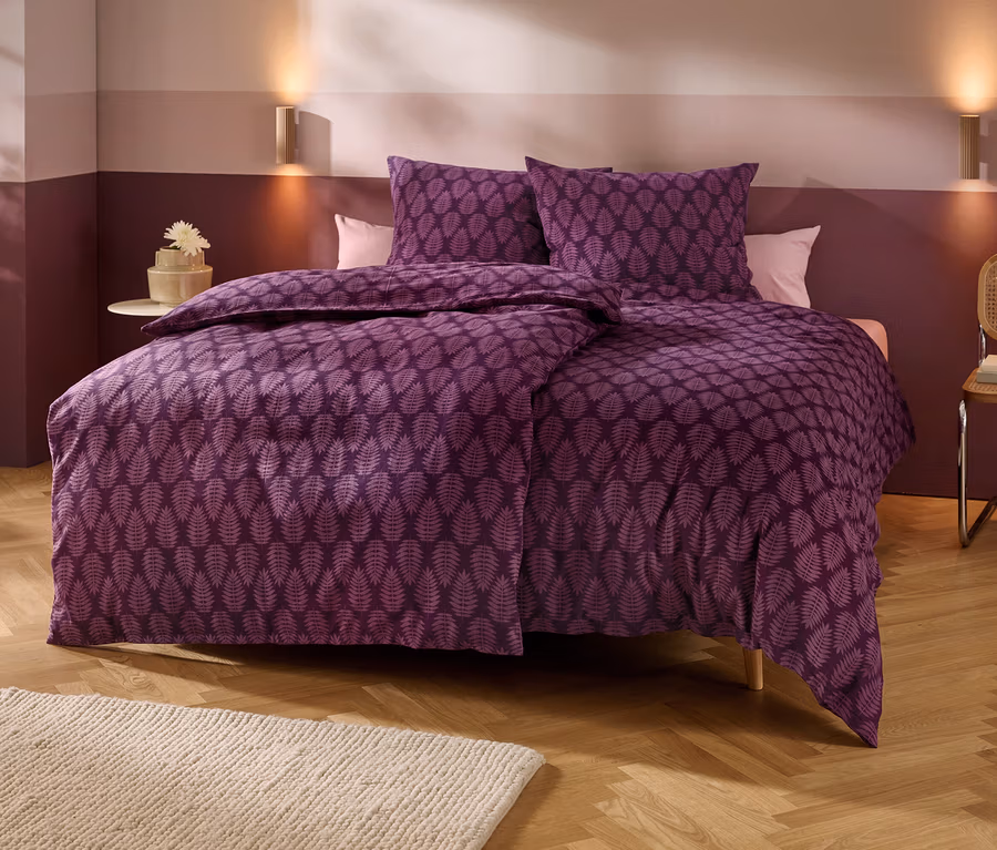 Ein Bett mit violetter Bettwäsche mit Blattmuster steht in einem Schlafzimmer mit zweifarbigen Wänden, Holzboden und einem hellen Teppich. Zwei Wandlampen beleuchten den Bereich über dem Bett. Links steht ein kleiner Beistelltisch mit einer Blume, rechts ein Stuhl mit Büchern.