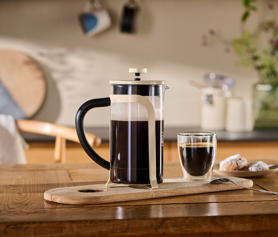 Eine Siebstempelkanne (800 ml, 6 Tassen) in friendly beige steht auf einem Holzbrett neben einer Tasse Kaffee.