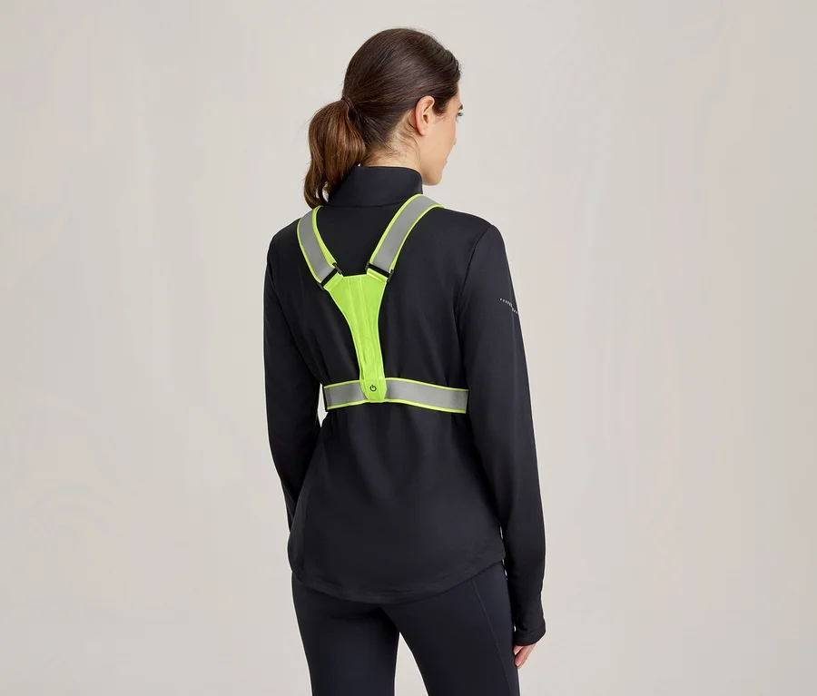 Frau von hinten, trägt ein schwarzes Langarmshirt und schwarze Leggings, darüber eine neongelbe Reflektorweste mit reflektierenden Streifen und einem kleinen Lichtknopf.