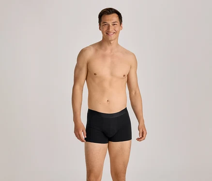 Oberkörperfreier Mann in schwarzen Boxershorts lächelt in die Kamera.