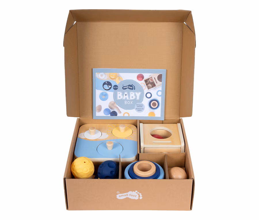 Offene small foot Baby-Box, 10–12 Monate, aus Karton, mit einer Broschüre und verschiedenen Holz- und Texturspielzeugen für Babys, darunter ein blau-gelber Formensortierer, eine Holzkiste mit Schublade, gelbe und blaue Texturbälle, blaue Stapelringe und ein Holzei.