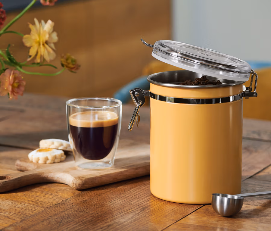 Kaffee in einem Glas, Kekse auf einem Holzbrett und ein offener gelber Kaffeebehälter mit Bohnen, dazu ein Messlöffel, auf einem Holztisch mit Blumen.