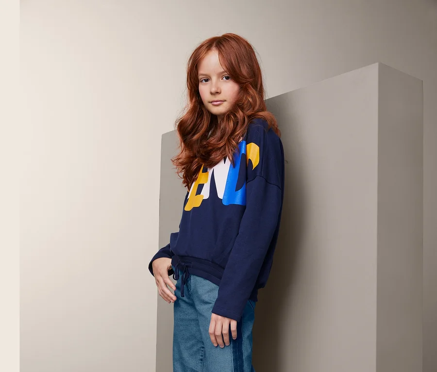 Ein junges Mädchen mit gewellten, roten Haaren posiert in einem dunkelblauen Sweatshirt, einer Jeans-Culotte mit Streifen und weißen Turnschuhen.