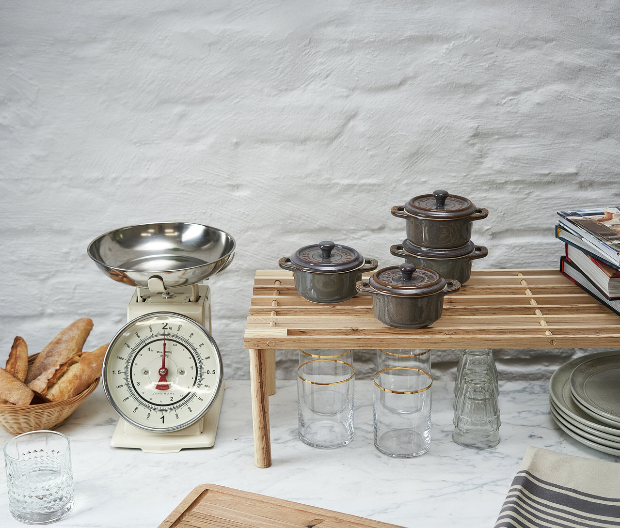 Vier antikgraue STAUB Keramik-Cocottes sind auf einem Holzregal auf einer Küchenarbeitsplatte angeordnet. Links daneben sind eine Vintage-Küchenwaage mit Silberschale und ein Brotkorb mit Baguettes zu sehen. Klare Gläser und gestapelte graue Teller befinden sich ebenfalls auf der Arbeitsplatte.