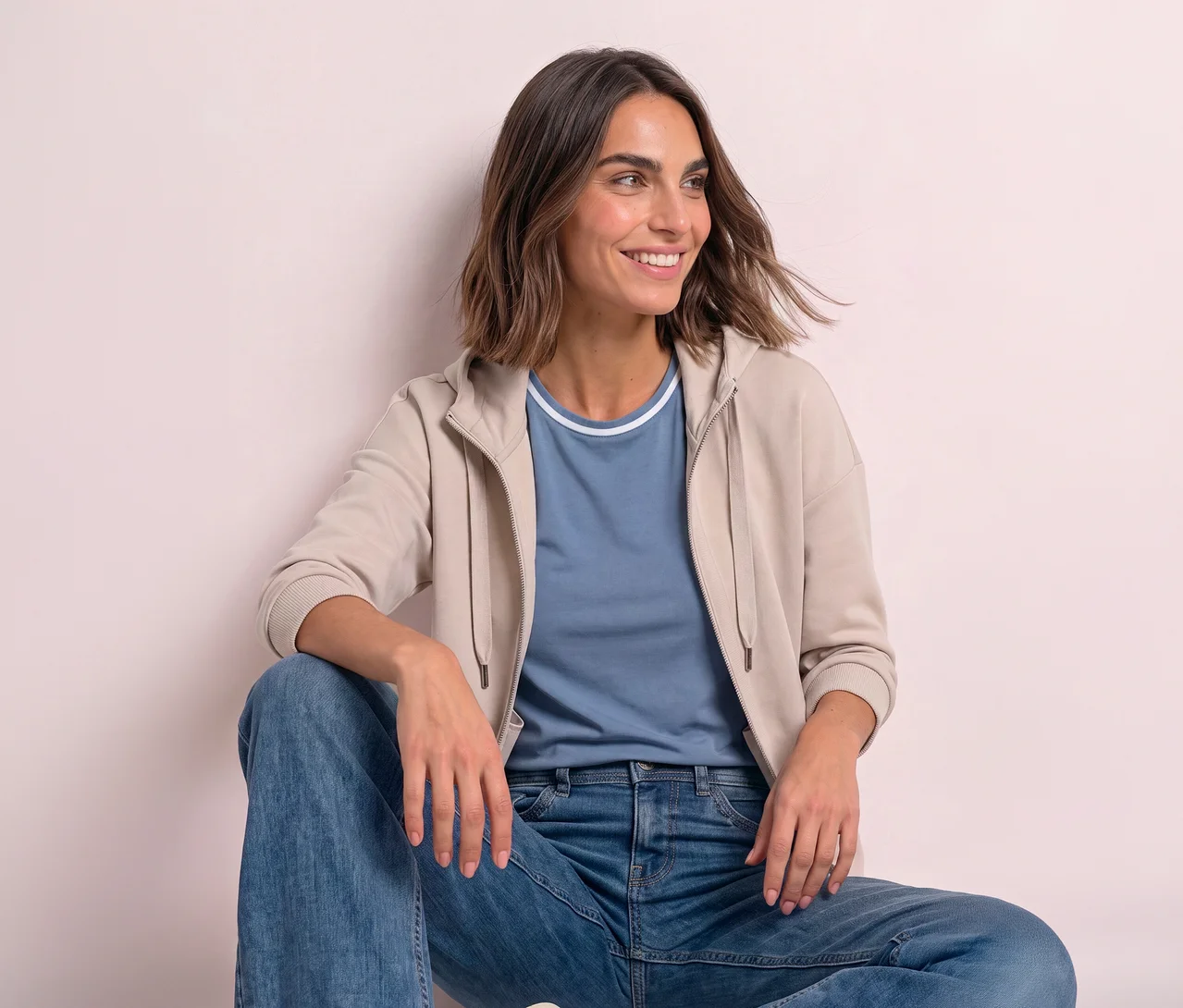 Lächelnde Frau sitzt, trägt ein hellblaues T-Shirt, blaue Jeans und eine offene beigefarbene Street One Sweatjacke.