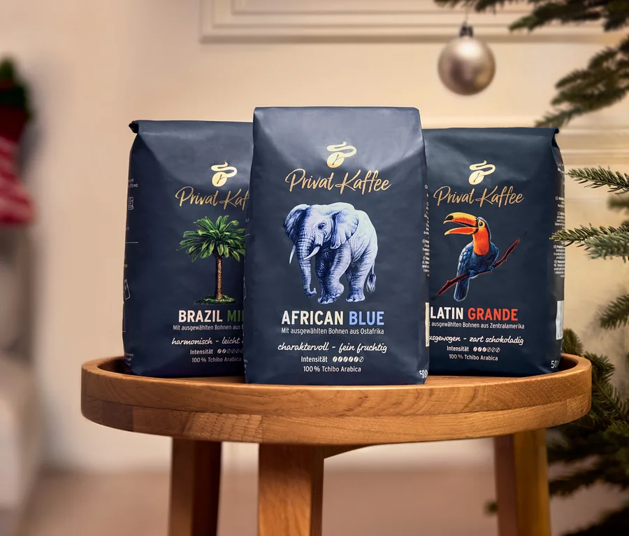 Drei 500g Packungen Tchibo Privat Kaffee Ganze Bohne stehen auf einem runden Holztisch vor einem weihnachtlich dekorierten Hintergrund. Von links nach rechts sind es Privat Kaffee Brazil Mild, Privat Kaffee African Blue und Privat Kaffee Latin Grande.