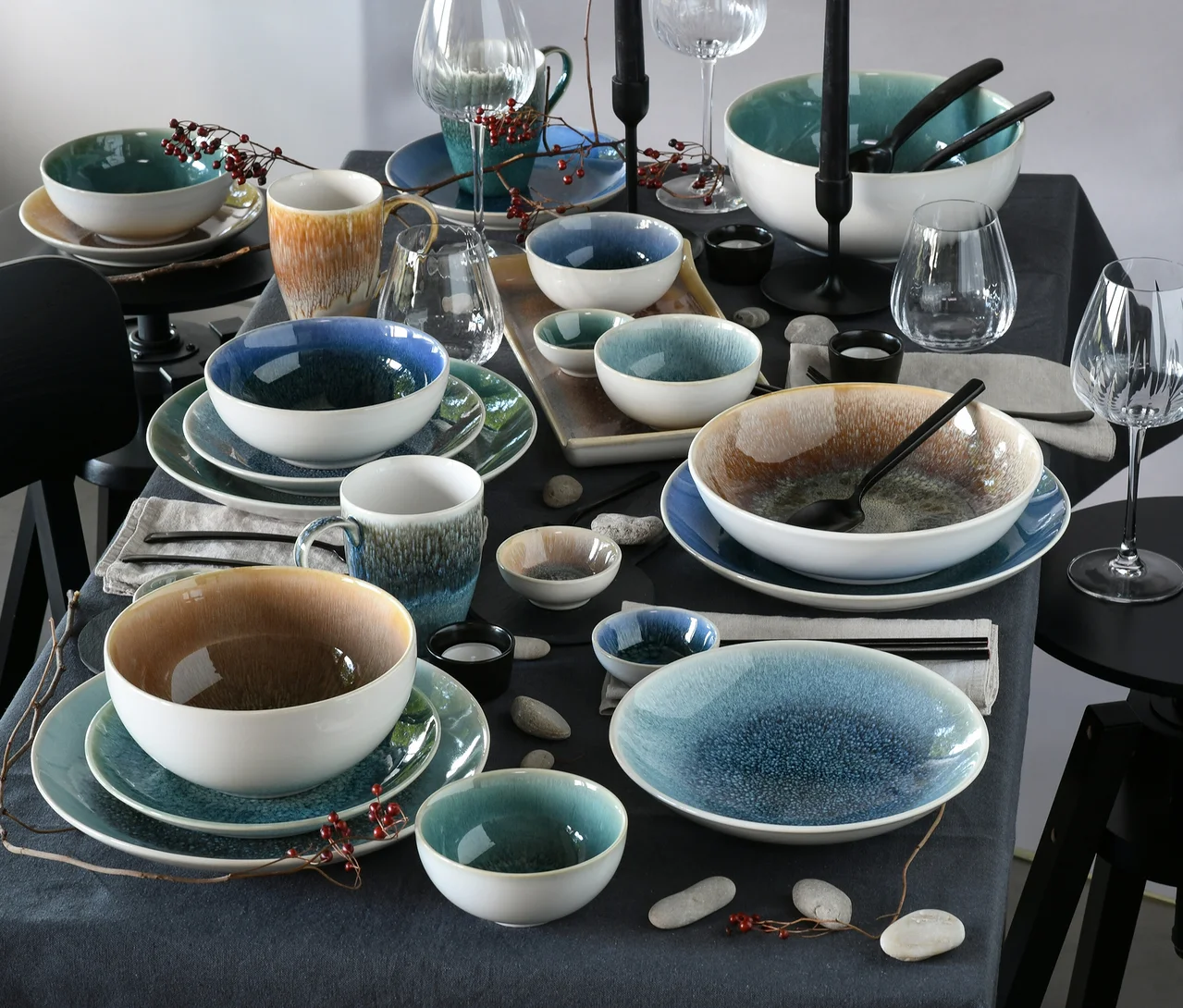 Ein gedeckter Tisch mit einem creaTable Bowl-Set »Caldera«, 12-teilig, bestehend aus Schalen, Tellern und Tassen in Blau-, Grün- und Brauntönen mit reaktiver Glasur, dekoriert mit Gläsern, Kerzen und Zweigen.