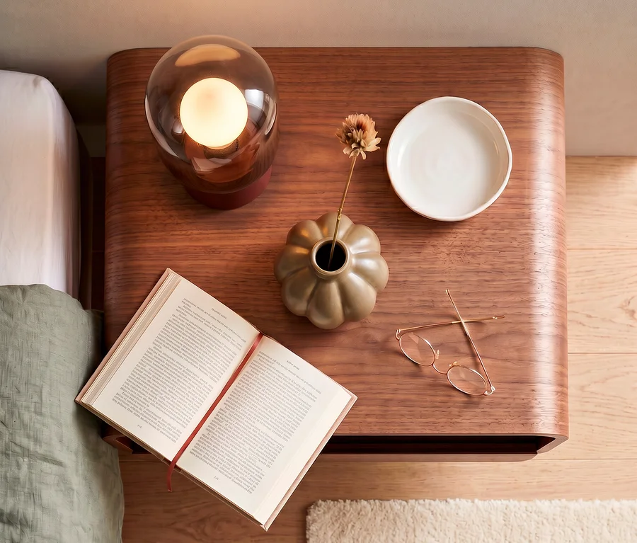 Draufsicht auf einen braunen WOOOD hängenden Holz-Nachttisch »Momo«. Darauf befinden sich eine leuchtende Lampe, eine beigefarbene kürbisförmige Vase mit einer Trockenblume, eine weiße Schale, ein offenes Buch und eine goldgerahmte Brille.