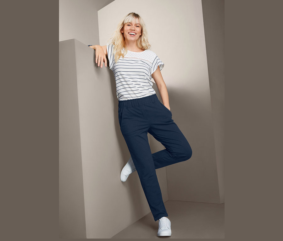 Blonde Frau lehnt an einer Wand und trägt ein gestreiftes T-Shirt und eine knöchellange Sweathose.