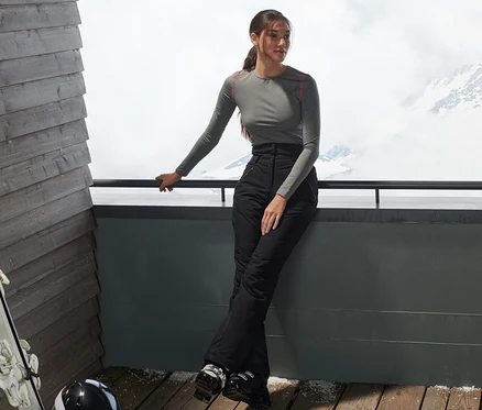 Frau lehnt an Geländer, trägt High-Waist-Skihose in Schwarz. Im Hintergrund schneebedeckte Berge.