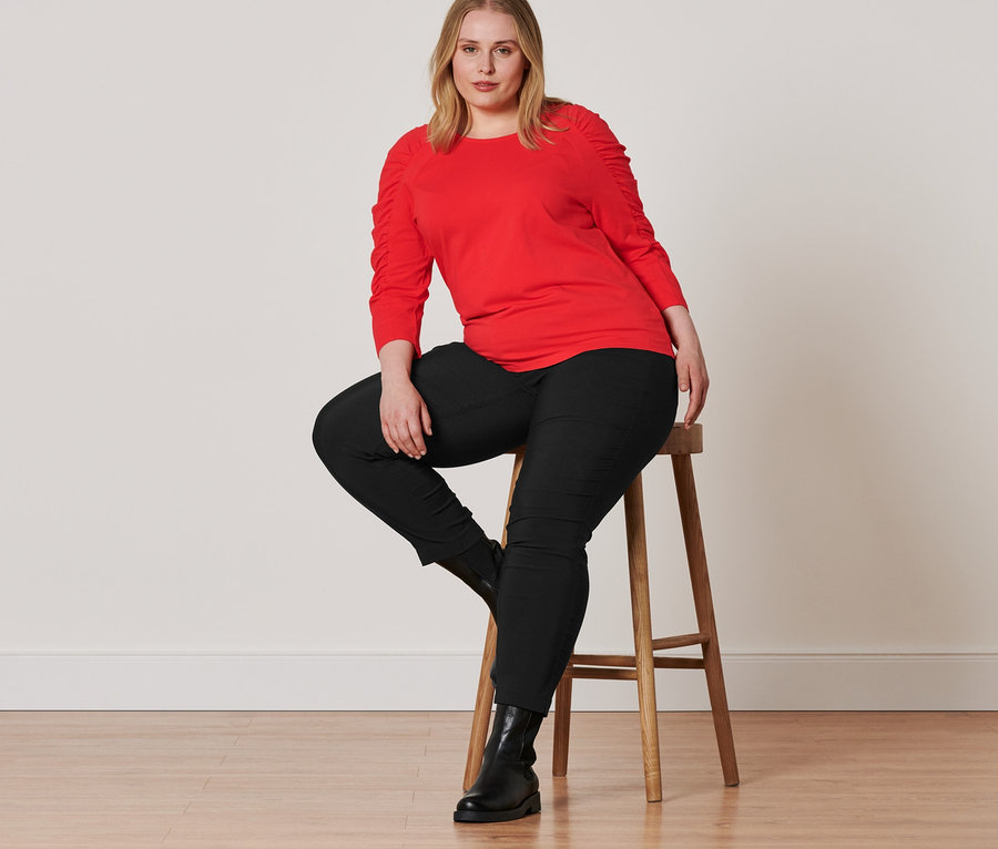 Eine Frau sitzt auf einem Hocker und trägt Lederboots, eine schwarze Bengalin-Hose und ein rotes Shirt mit 3/4-Arm.