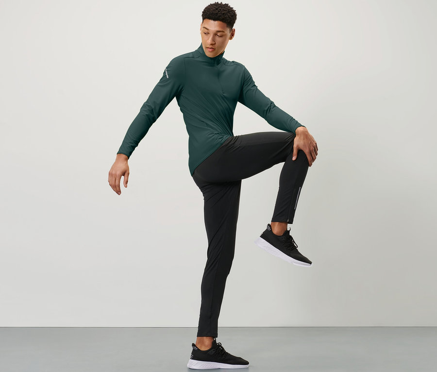 Mann in grünem Sportoberteil und schwarzen Windprotection-Lauftight hält ein Knie mit einer Hand vor der Brust.