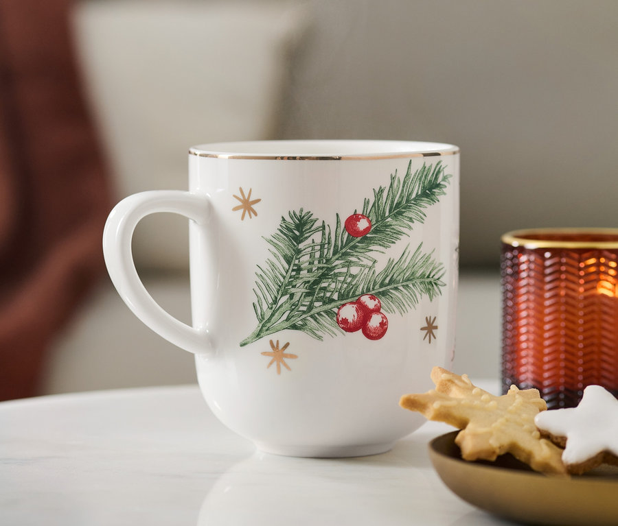 Nahaufnahme eines Jumbobechers mit Glanzgolddekor und Weihnachtsmotiv, der neben einem Teller mit Keksen und einer Kerze steht.