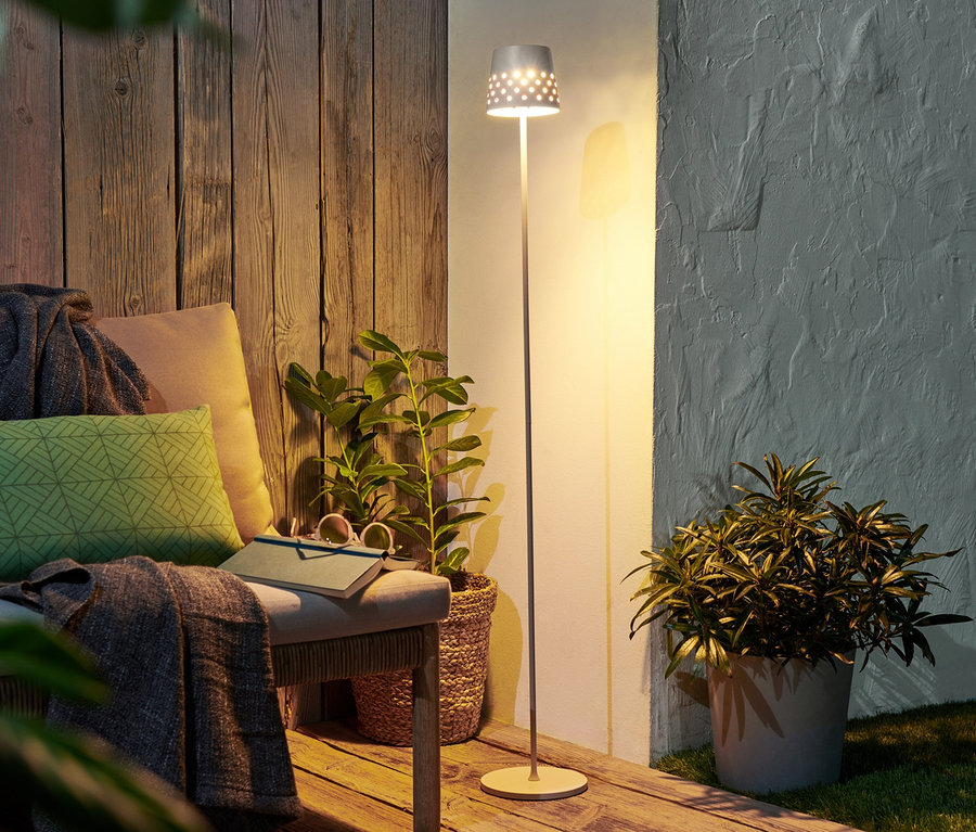 Eine LED-Akku-Stehleuchte aus Metall steht neben einer Bank mit Kissen und einem Buch.