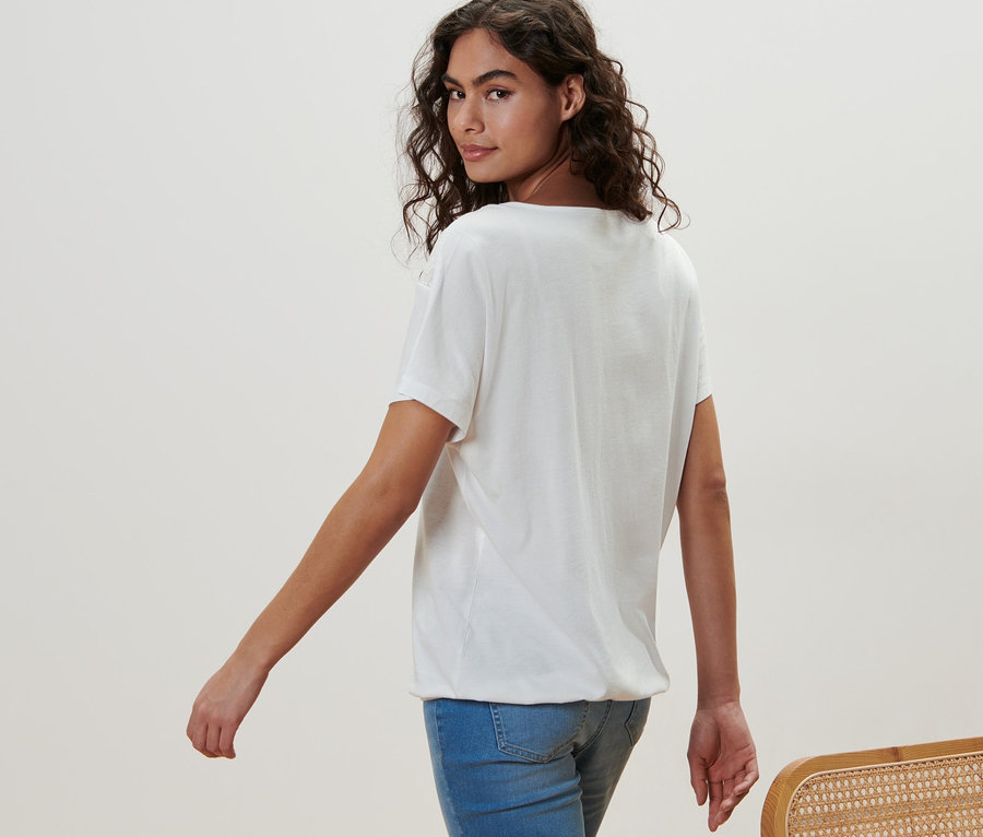 Frau geht und schaut über die Schulter. Sie trägt ein weißes Blusenshirt mit Stickerei und Jeans.