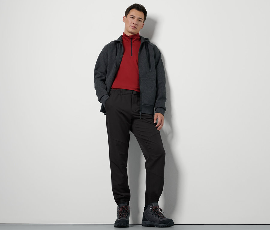 Ein Mann steht und trägt eine Thermohose mit Gürtel, eine Zip-Jacke und Wanderschuhe.