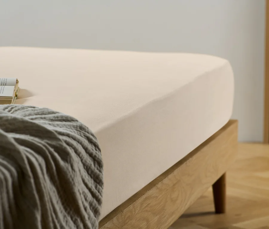 Ein beiges Spannbettlaken auf einem Bett mit Holzrahmen. Auf dem Bett liegen ein Buch und eine graue Decke.