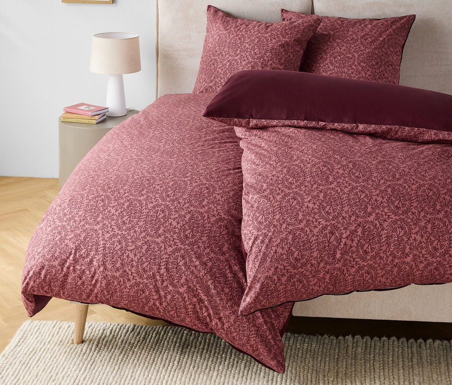 Bettwäsche mit Blumenmuster in Rosa- und Burgundertönen auf einem Bett mit beigem Kopfteil.