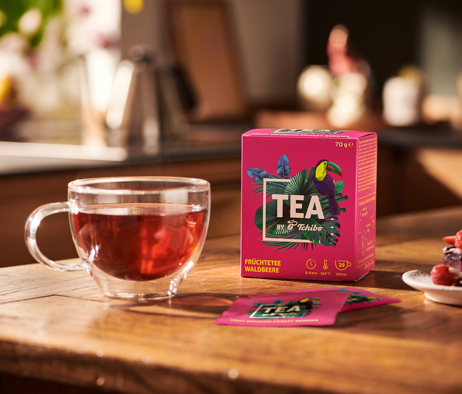 Auf einem Holztisch stehen eine Glastasse mit Früchtetee, eine Packung TEA by Tchibo Früchtetee Waldbeere und ein Teller mit Dessert.