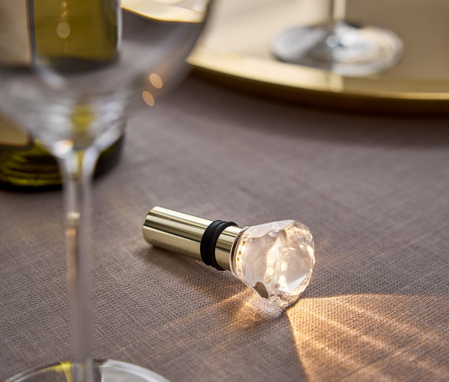 Auf einem grauen Tuch liegen ein goldener Weinstopfen mit Kristallkopf, daneben ein Glas und eine Flasche Wein.