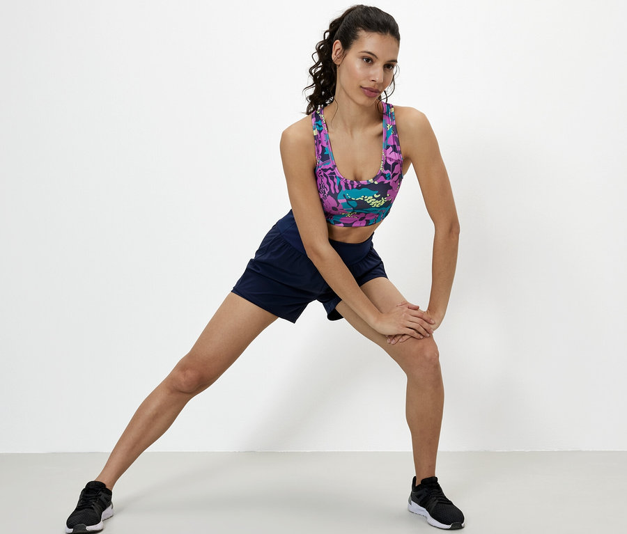 Frau dehnt Bein, trägt Sport-Bustier, Shorts und Schuhe.