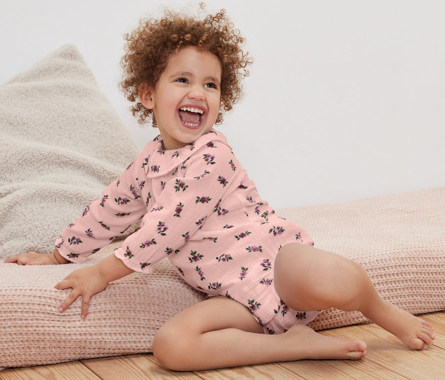 Ein lächelndes, lockiges kleines Mädchen sitzt auf einer Unterlage und trägt einen Baby-Musselin-Body mit Blumenmuster.