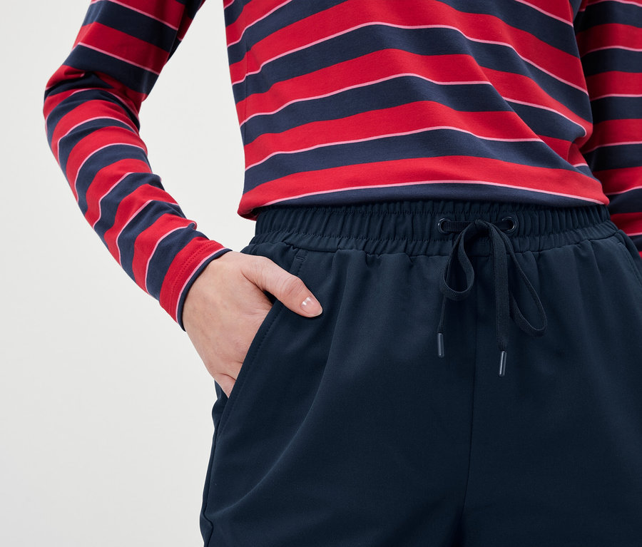 Detailaufnahme einer Frau, die marineblaue Trainingshose und ein rot-blau gestreiftes Langarmshirt trägt und eine Hand in der Hosentasche hat.
