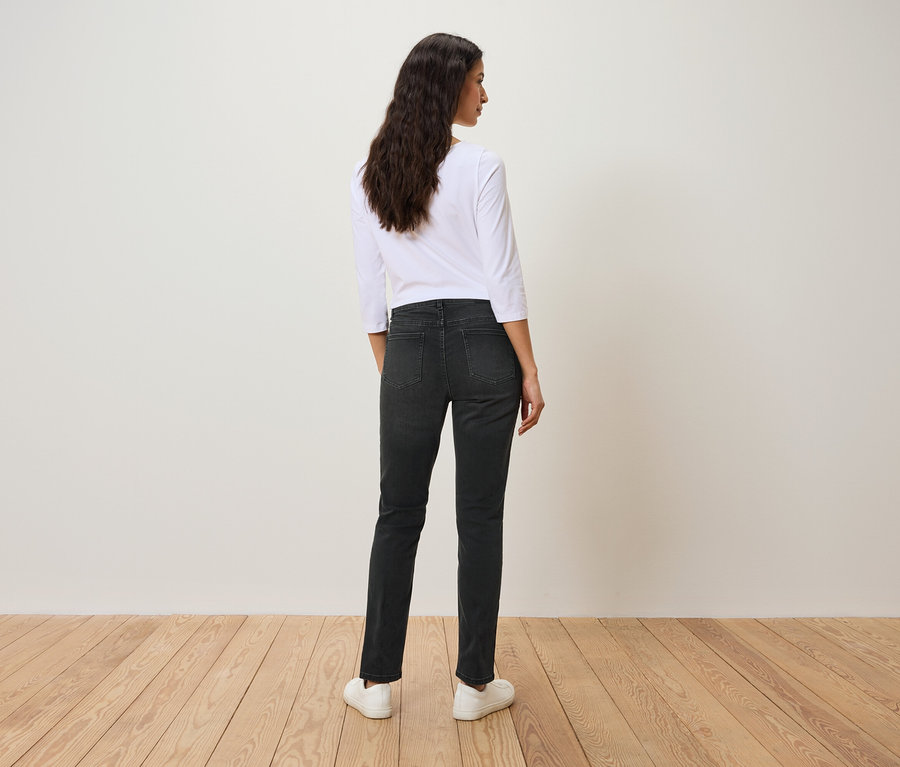 Rückansicht einer Frau mit dunklem, gewelltem Haar, die ein weißes Dreiviertelarm-Shirt, schwarze Jeans und weiße Schuhe trägt.