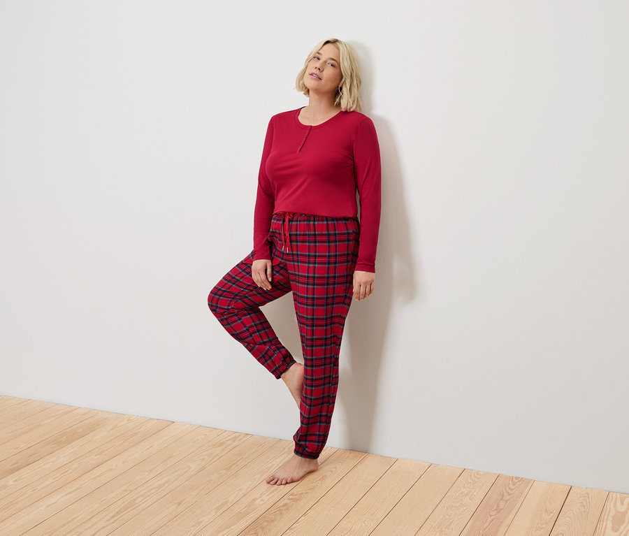 Blonde Frau in rotem Pyjama mit karierter Hose steht an einer weißen Wand.