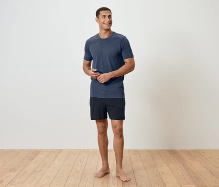 Mann steht barfuß auf einem Holzboden, trägt ein blaues T-Shirt und schwarze Shorts.