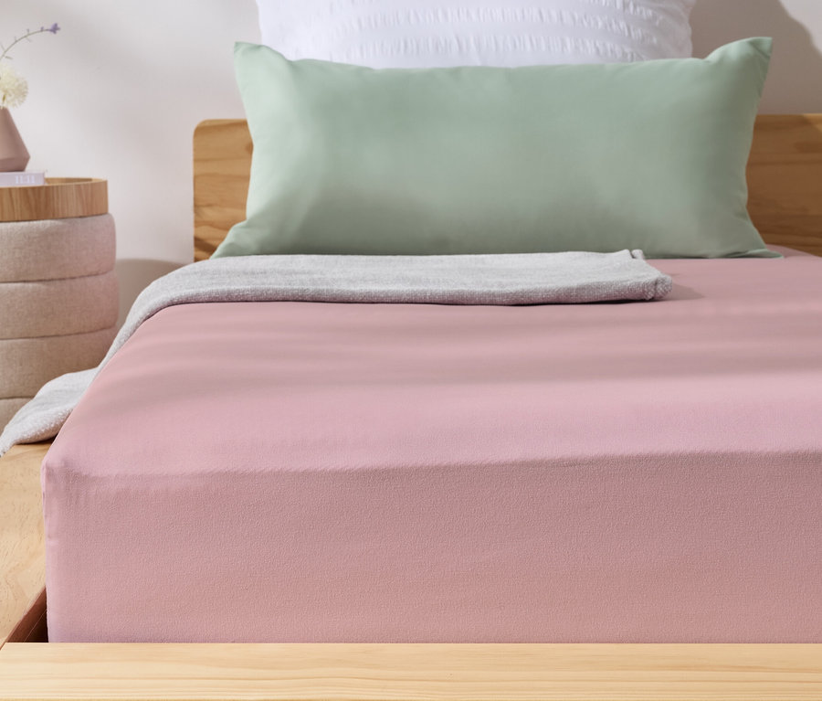 Rosa Microfaser-Spannbettlaken auf einem Bett. Daneben steht ein Nachttisch mit einer Vase.