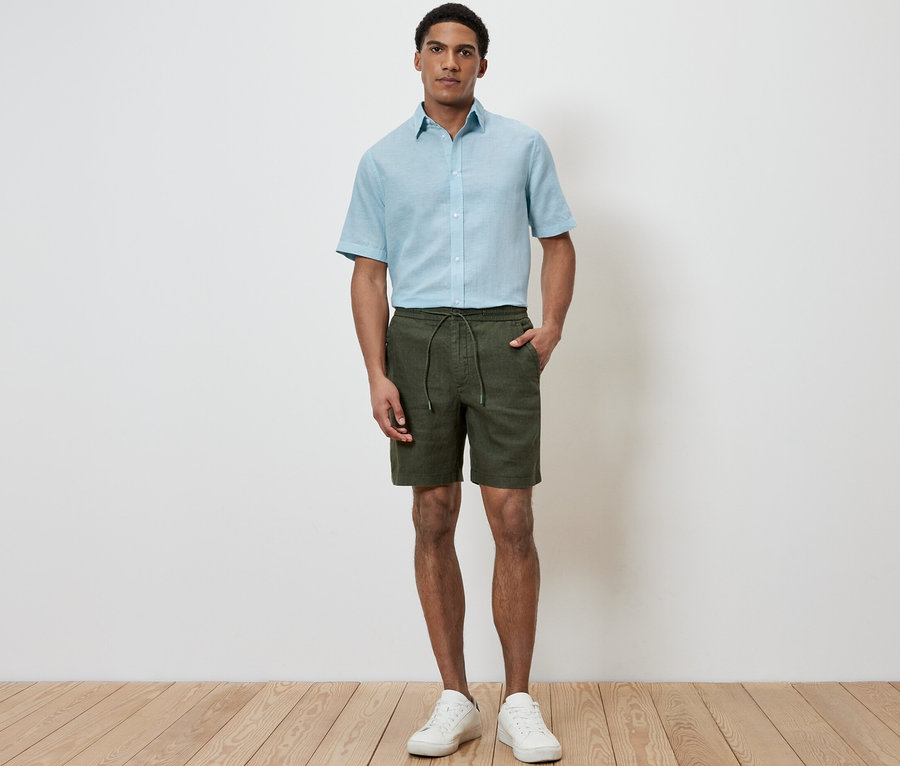Mann steht und trägt Leinen-Shorts in Khaki, hellblaues Hemd und weiße Turnschuhe.
