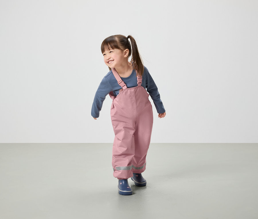 Lächelndes kleines Mädchen trägt eine rosafarbene Latzhose und blaue Gummistiefel.