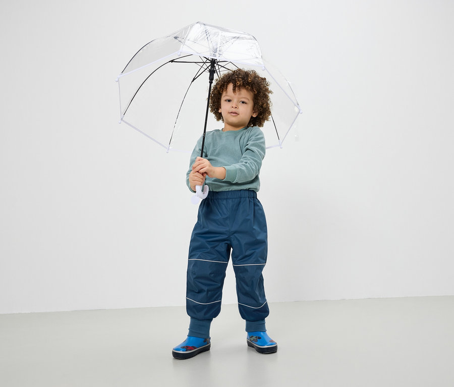 Ein Junge hält einen transparenten Regenschirm, trägt blaue Hosen und Gummistiefel.