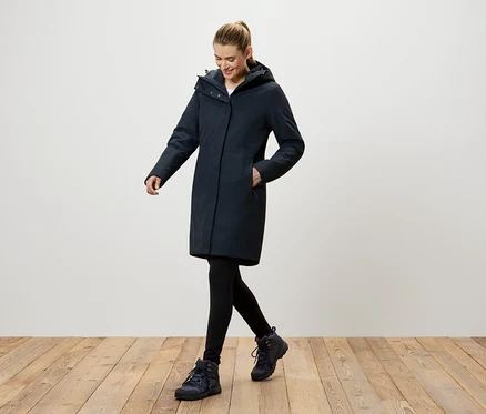 Eine Frau geht auf einem Holzboden und trägt einen dunkelblauen Mantel mit Kapuze, schwarze Leggings und dunkelblaue Schuhe.