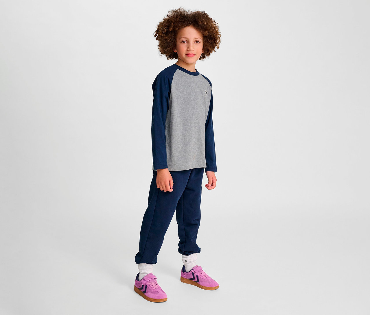 Junge steht. Er trägt blaue Trainingshose und graues HMLJR Raglan Loose T-Shirt BEE mit blauen Ärmeln, dazu rosa Schuhe.