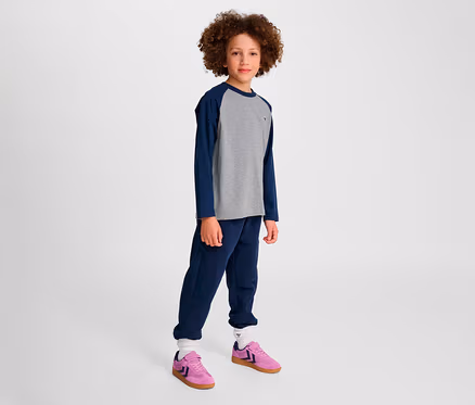 Junge steht. Er trägt blaue Trainingshose und graues HMLJR Raglan Loose T-Shirt BEE mit blauen Ärmeln, dazu rosa Schuhe.