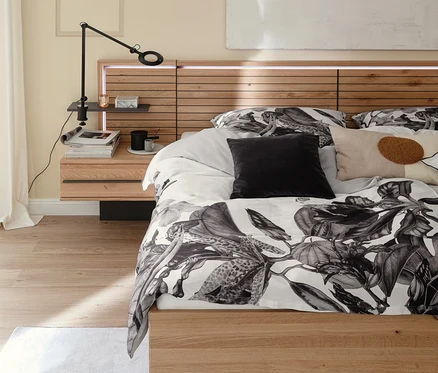 Bett mit Holzrahmen und beleuchtetem Kopfteil. Graue SCHÖNER WOHNEN-Kollektion Bettwäsche »AMALIA« mit Blättermuster und Kissen. Nachttisch mit Lampe.
